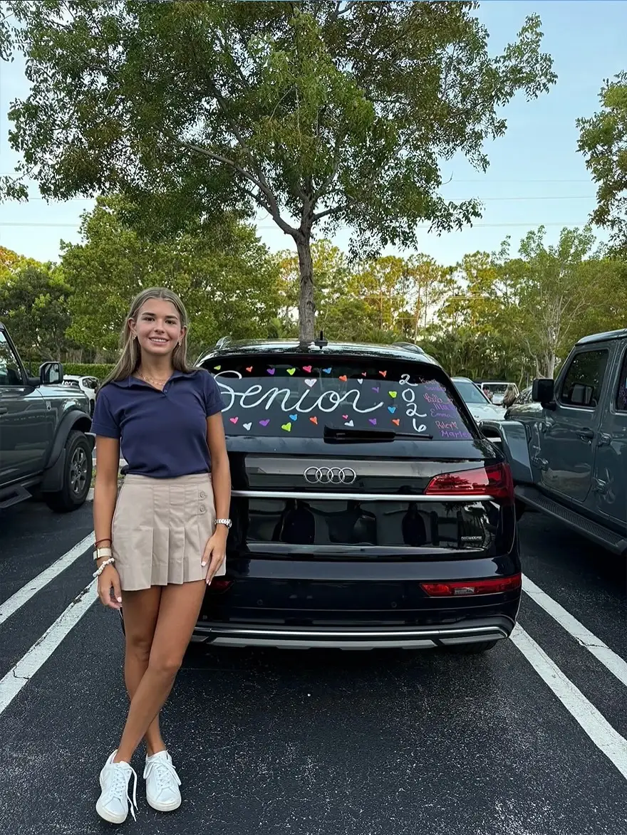Kai Trump stands smiling in a parking lot next to a black Audi SUV decorated with "Senior 2026" on the back window.
