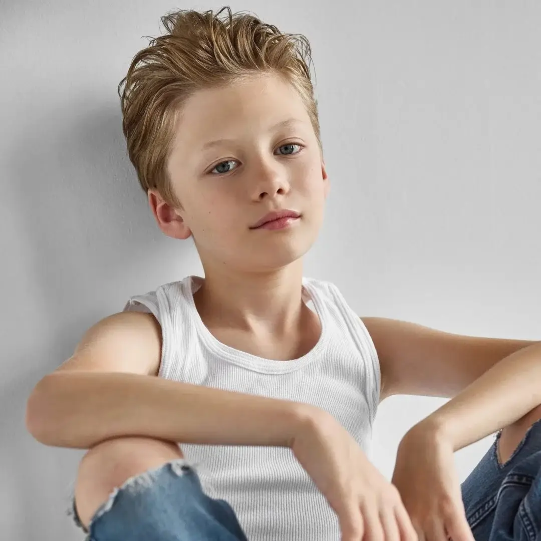 Theo Clark Leber with styled blonde hair sits leaning against a plain wall wearing a white tank top and ripped jeans.