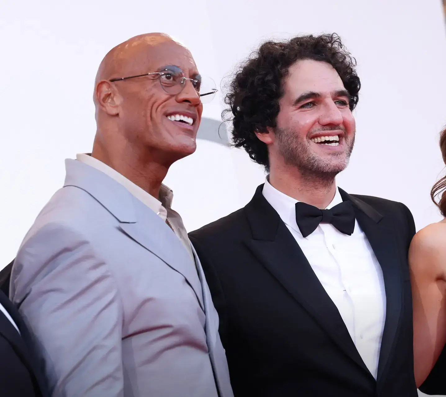 Dwayne Johnson in a grey suit and round glasses stands smiling alongside Benny Safdie in a classic black tuxedo.