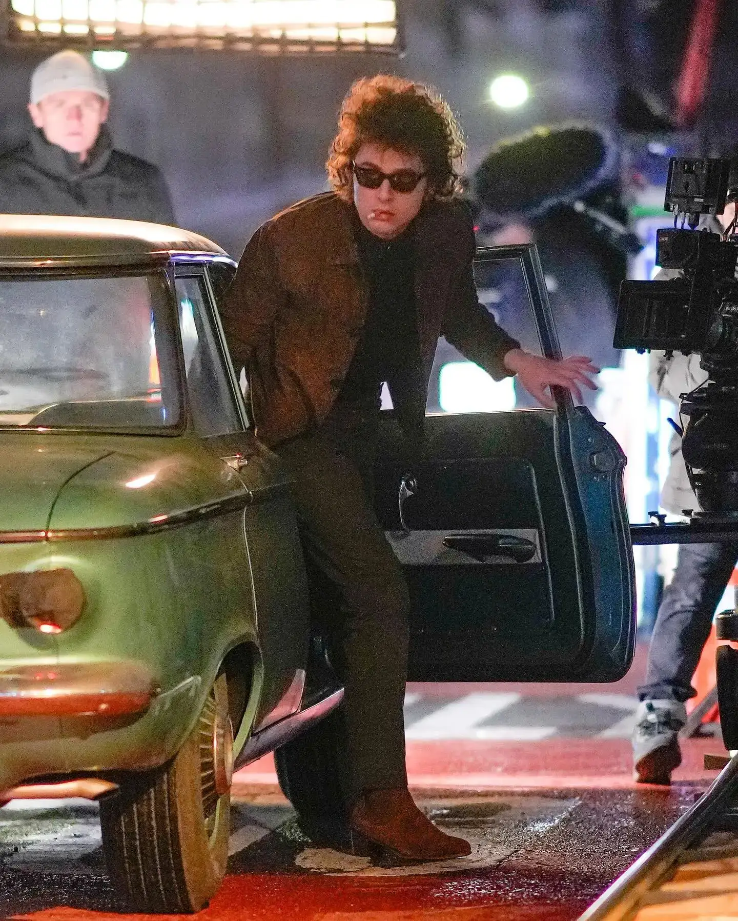 Timothée Chalamet as Bob Dylan wears sunglasses and a brown jacket with a cigarette in his mouth while stepping out of a vintage green car on set.