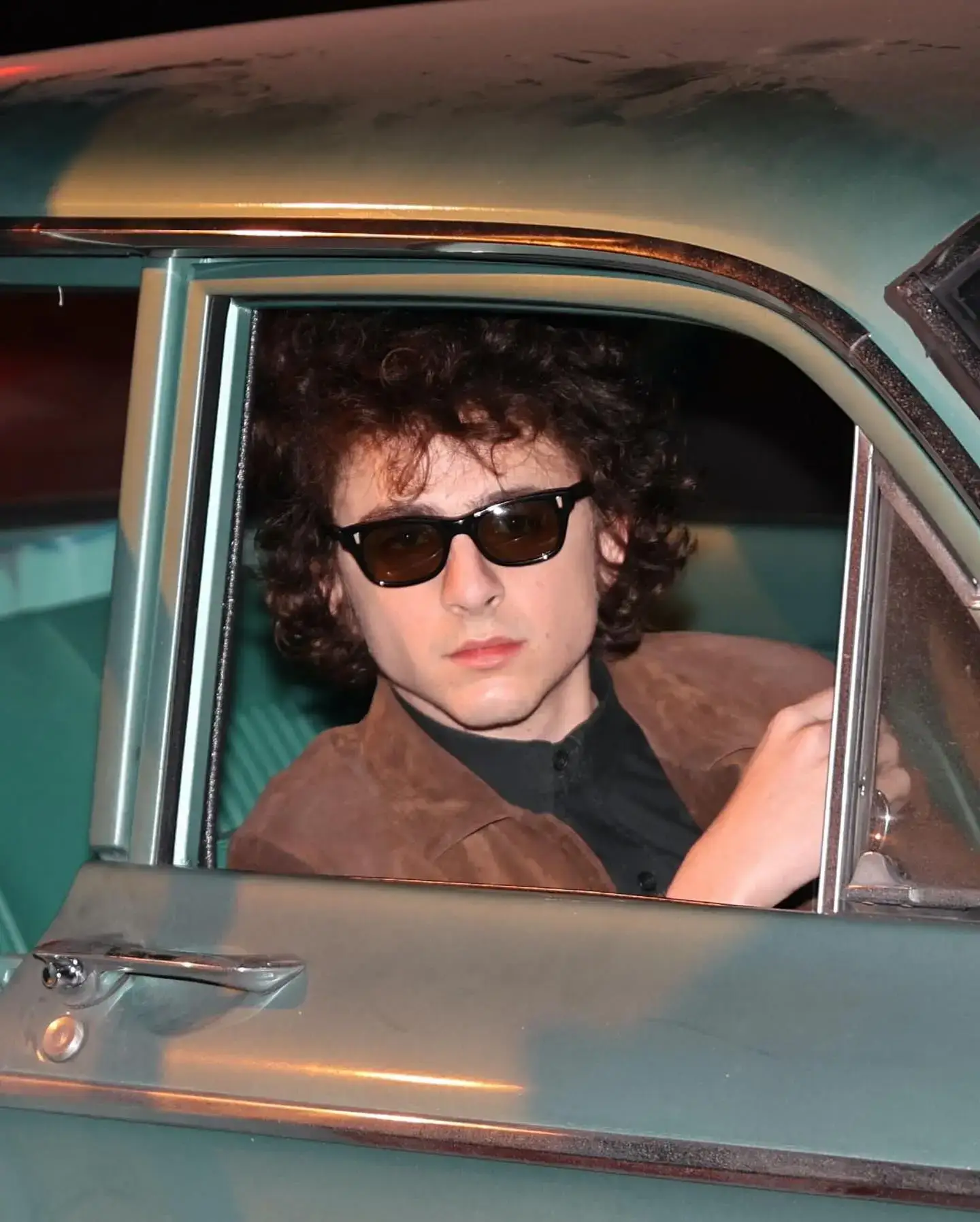 Timothée Chalamet as Bob Dylan wears sunglasses and a brown jacket while sitting in the driver