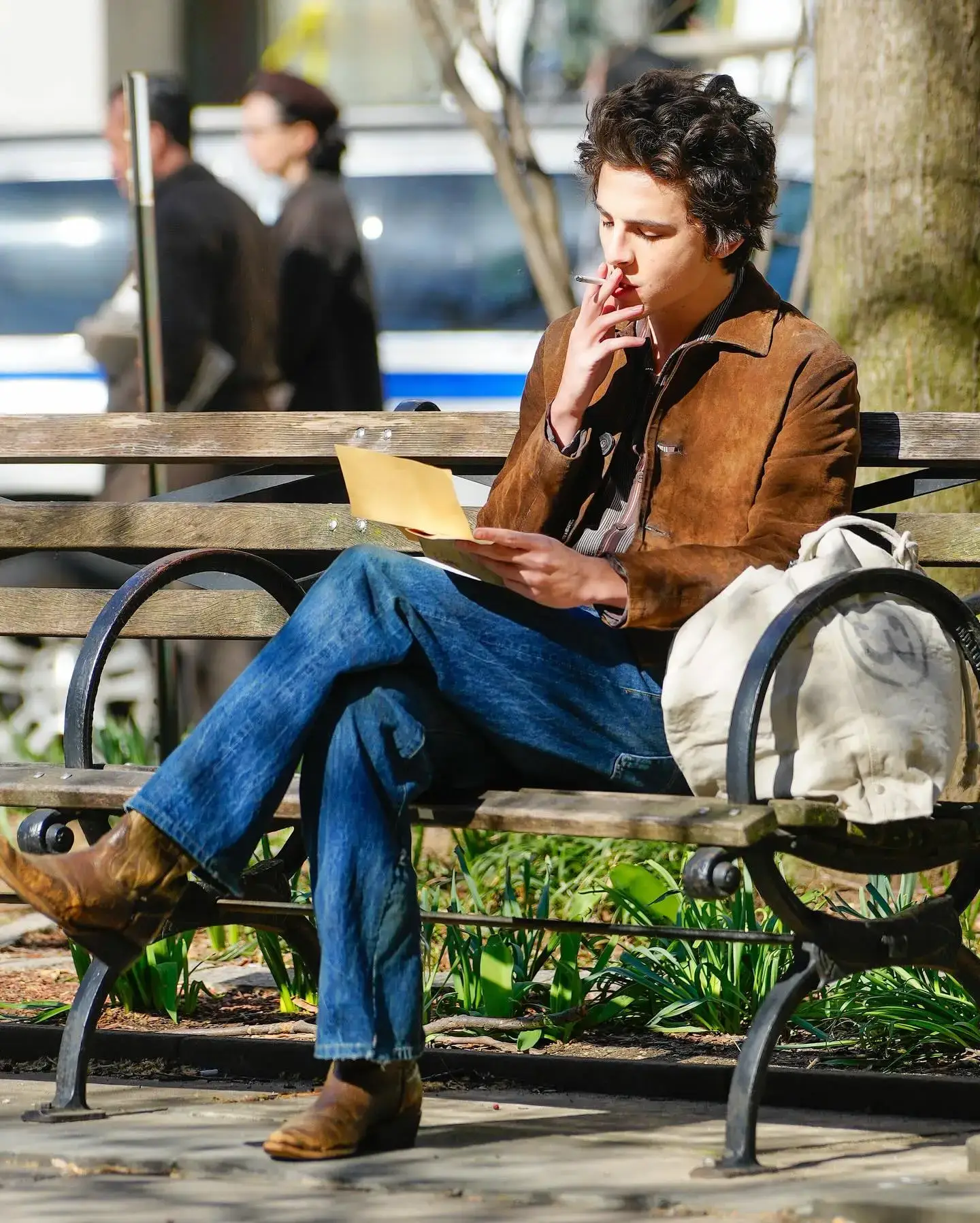 Timothée Chalamet sits cross-legged on a park bench wearing a brown suede jacket and jeans while reading a paper and holding a cigarette.