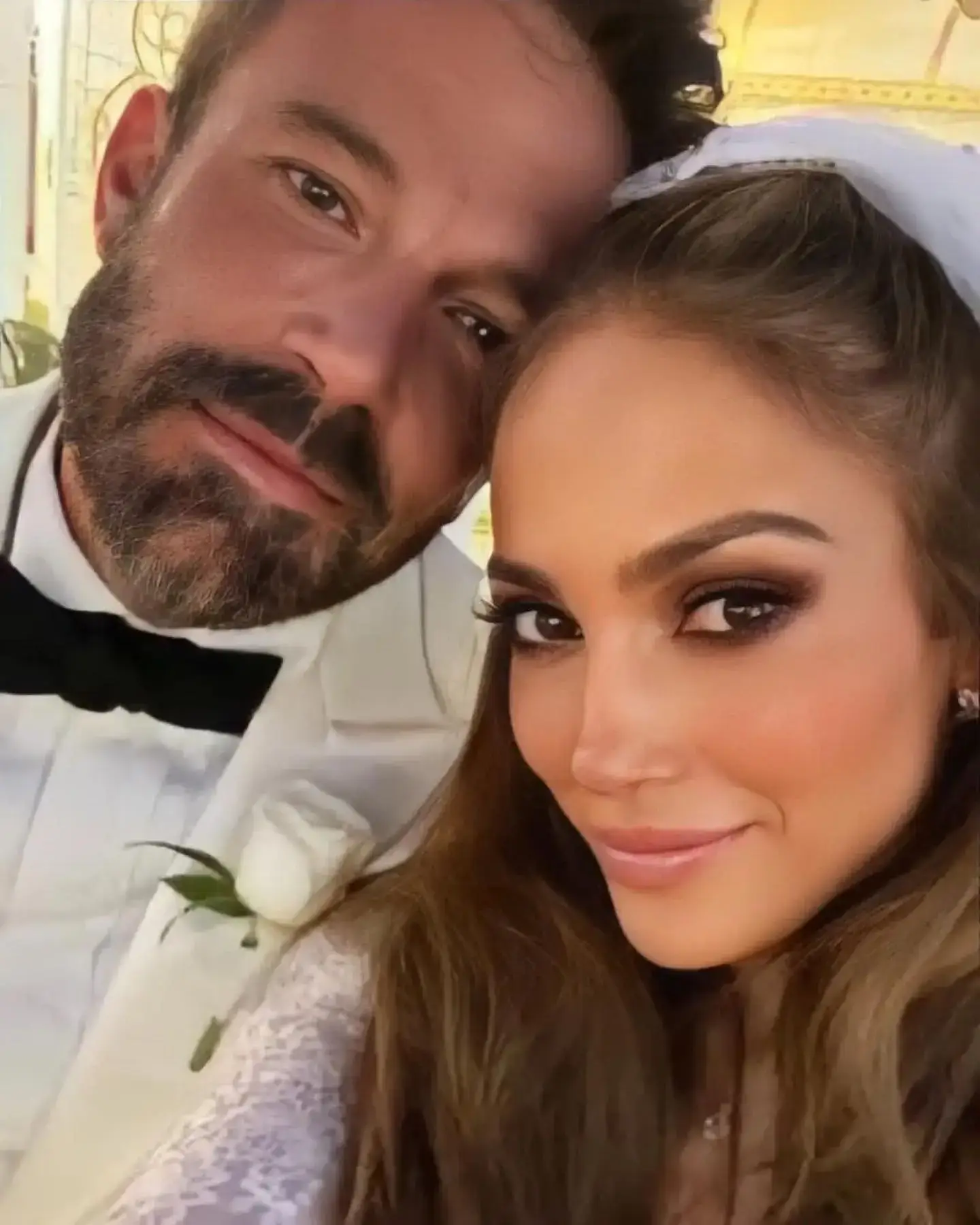 Jennifer Lopez in a wedding veil smiling next to Ben Affleck in a white tuxedo jacket and black bow tie.