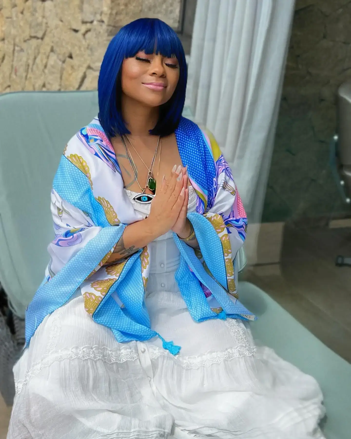 Singer Nivea sits serenely with her hands in prayer position, wearing a blue bob wig, a white dress, and a patterned shawl.