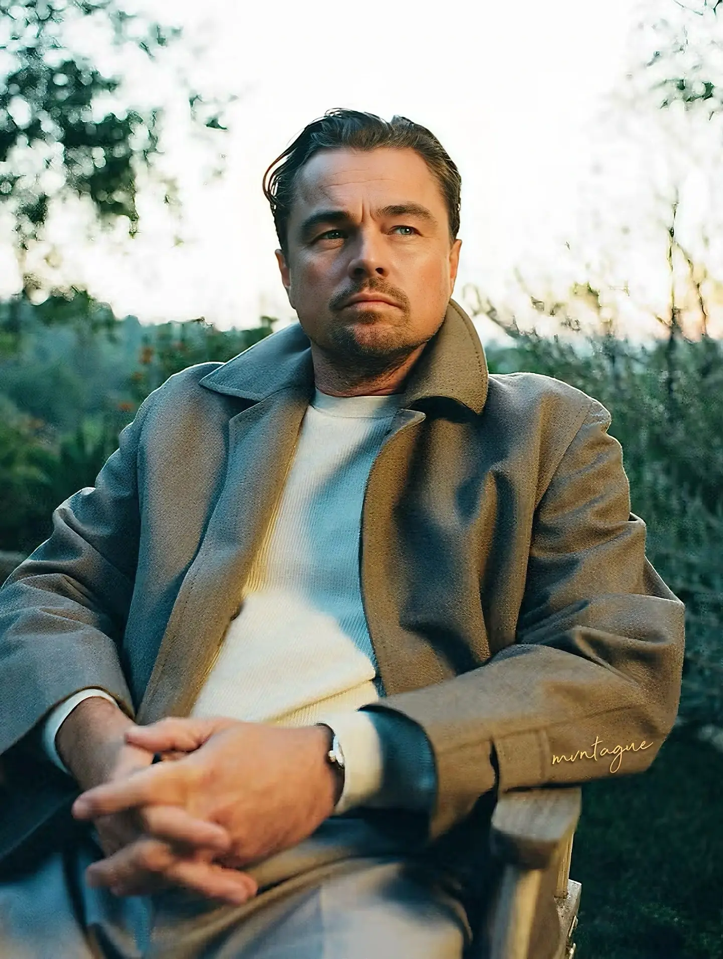 Actor Leonardo DiCaprio sits outdoors in a wooden chair wearing a grey jacket over a white sweater.