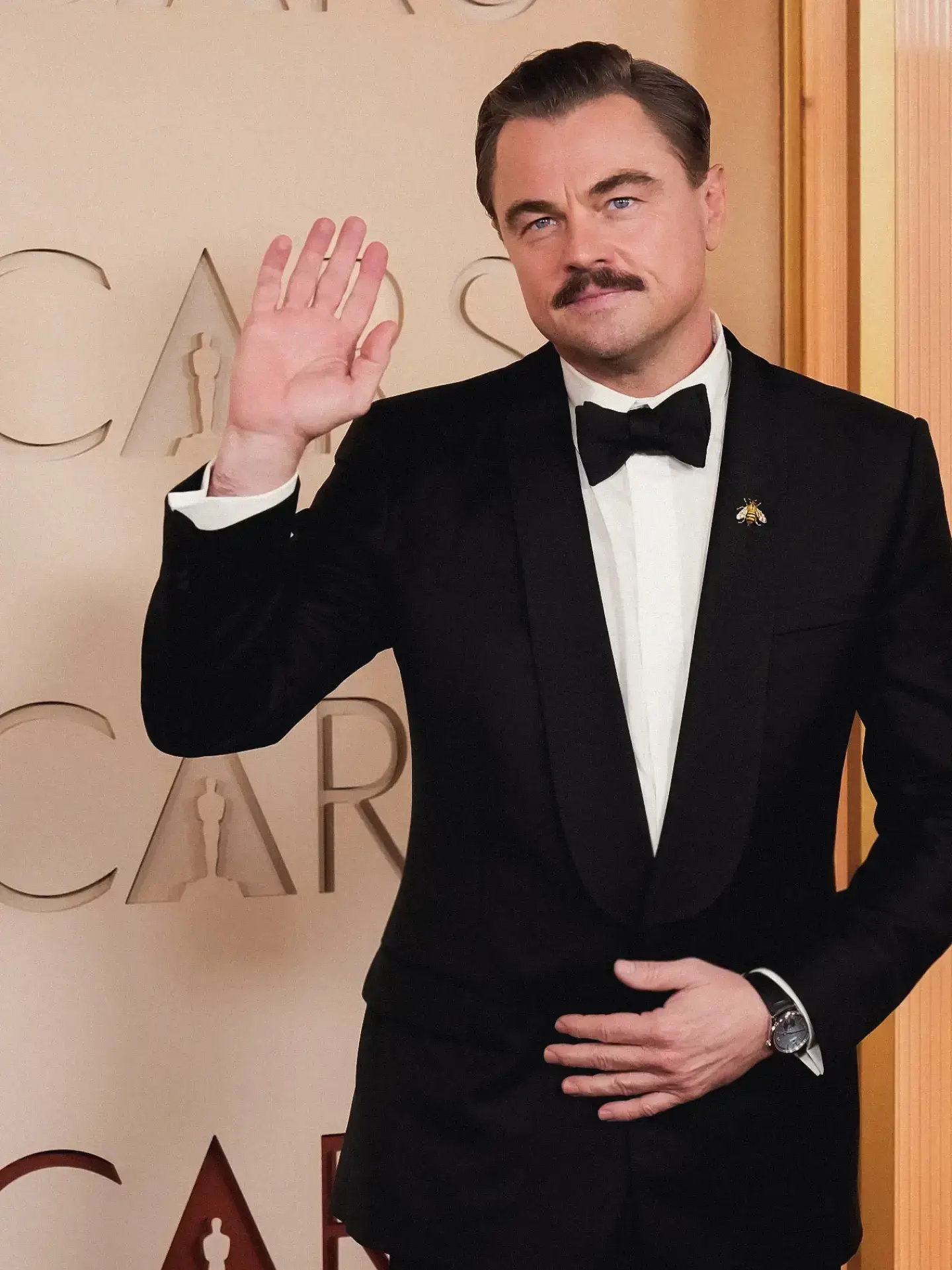Actor Leonardo DiCaprio sporting a mustache waves hand while wearing a black tuxedo with a bee lapel pin at an Oscars event.
