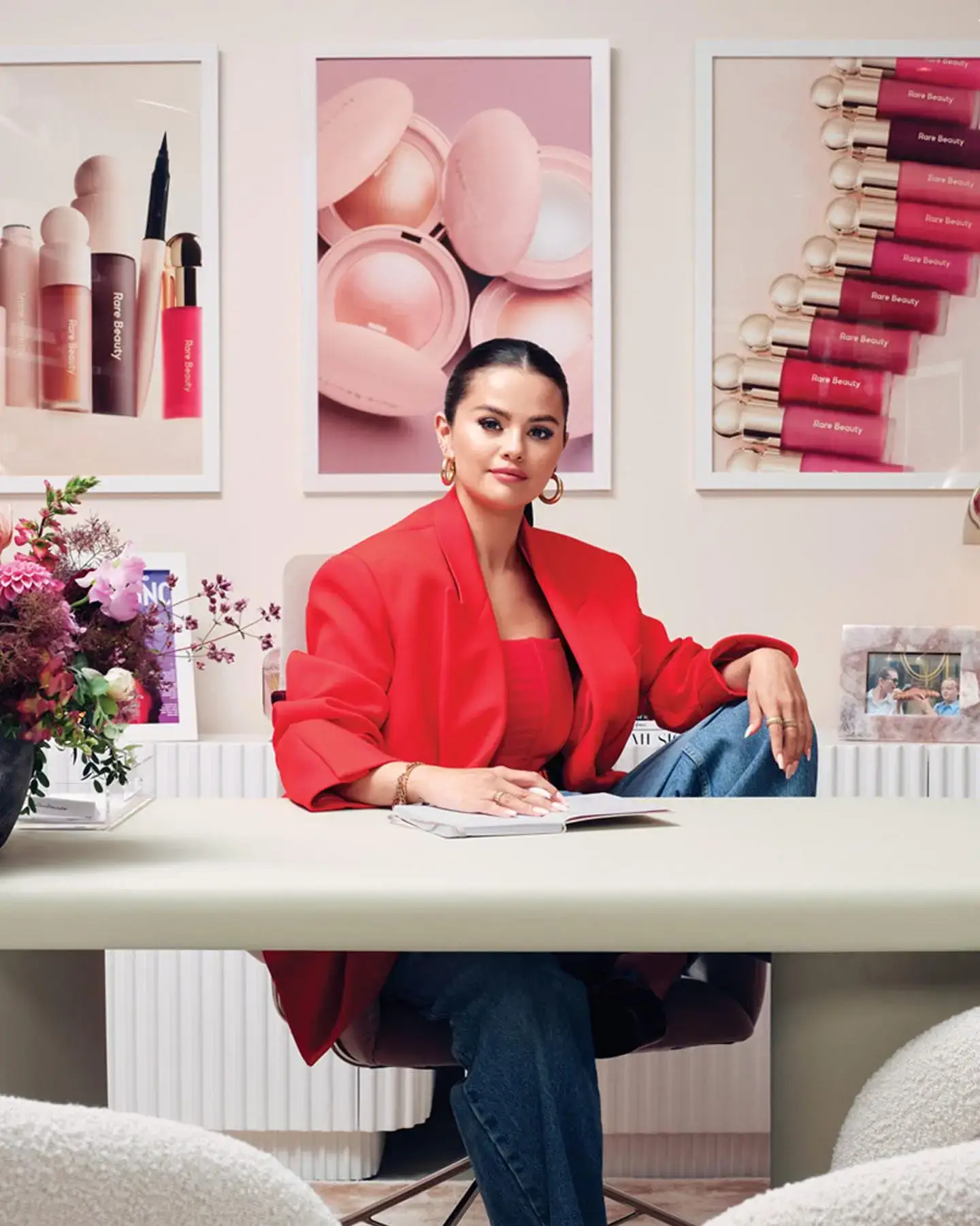 Selena Gomez wearing a bright red blazer sits at an office desk framed by Rare Beauty product posters.