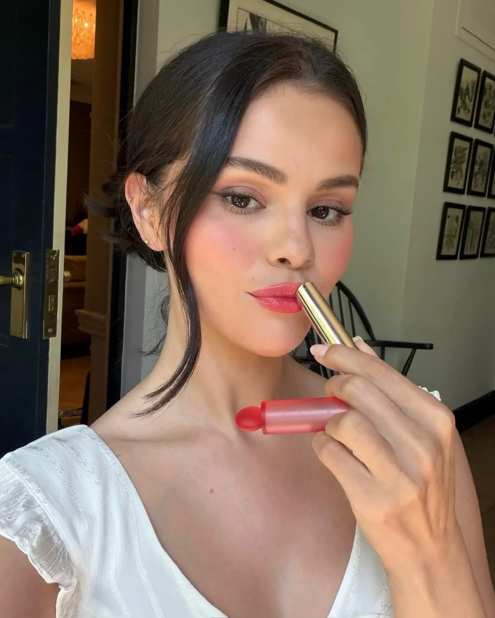 Selena Gomez applies a pink lip product to her lips while posing for a close-up selfie in a white top.