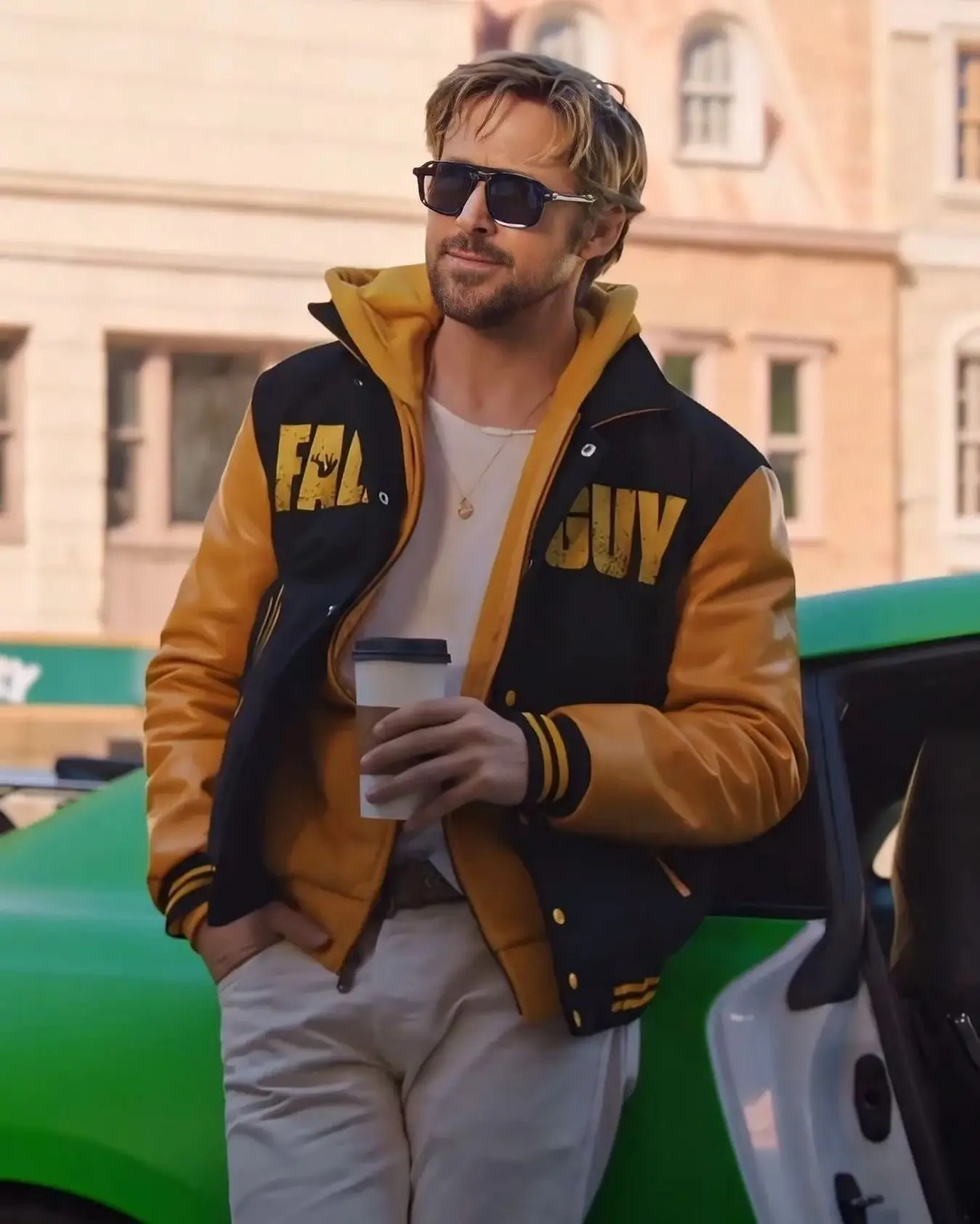 Ryan Gosling wears sunglasses and a yellow and black "Fall Guy" jacket while holding a coffee cup next to a green sports car.