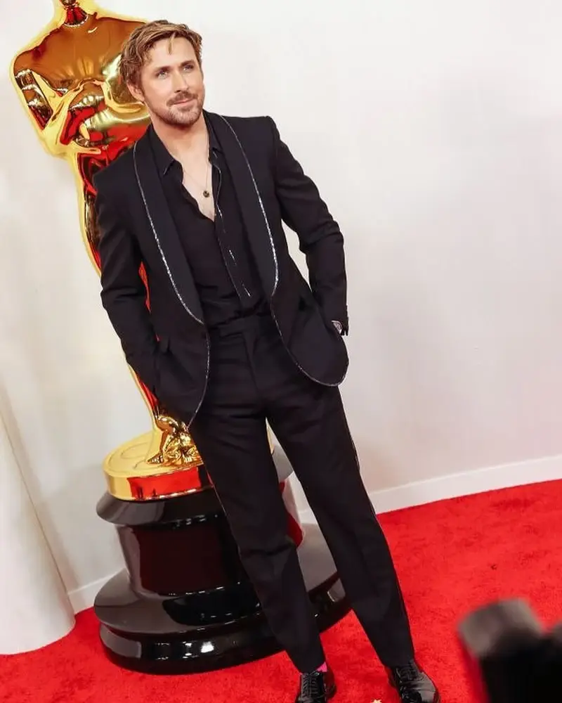 Actor Ryan Gosling dressed in a black suit with silver trim poses on a red carpet next to a large golden Oscar statue.