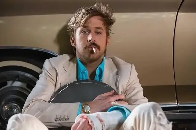 Actor Ryan Gosling with messily styled hair and a mustache sits against a car holding a hubcap with a cigarette in his mouth.