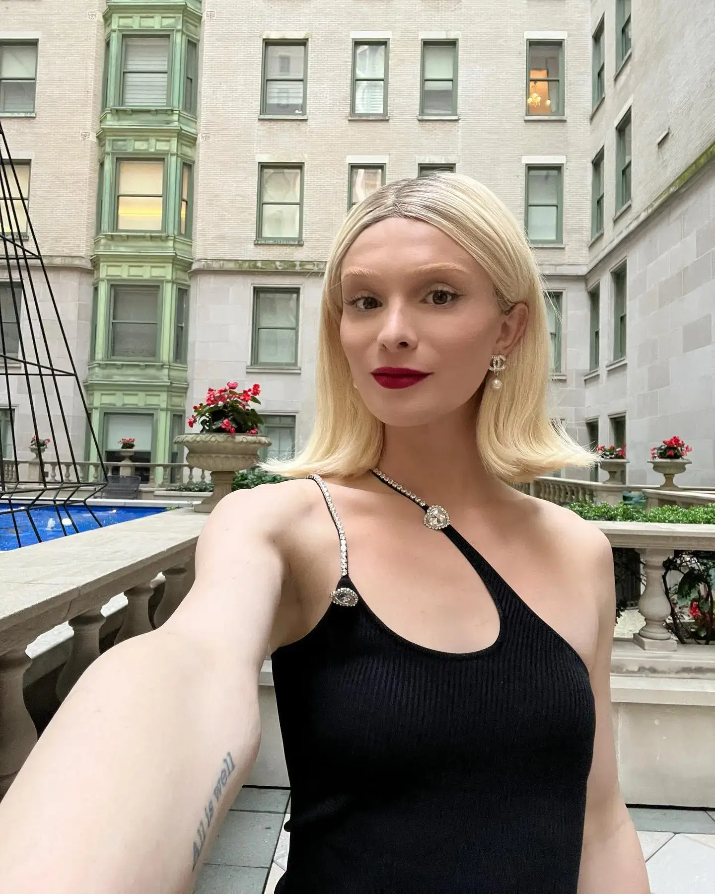 Content creator Dylan Mulvaney takes a selfie wearing a blonde bob wig and a chic black one-shoulder dress in an outdoor courtyard.