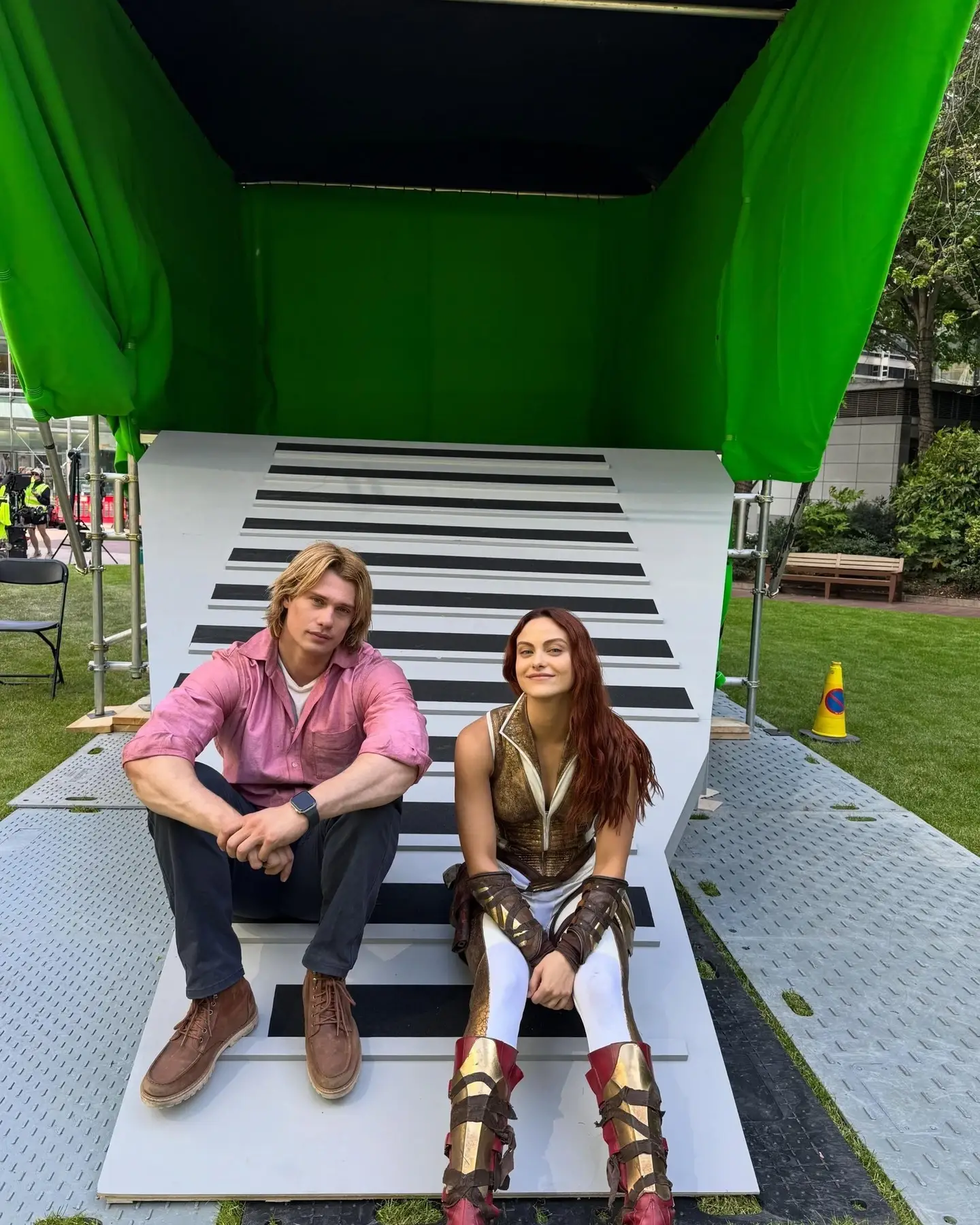 Actors Nicholas Galitzine and Camila Mendes sit together on a striped set ramp in front of a green screen structure.