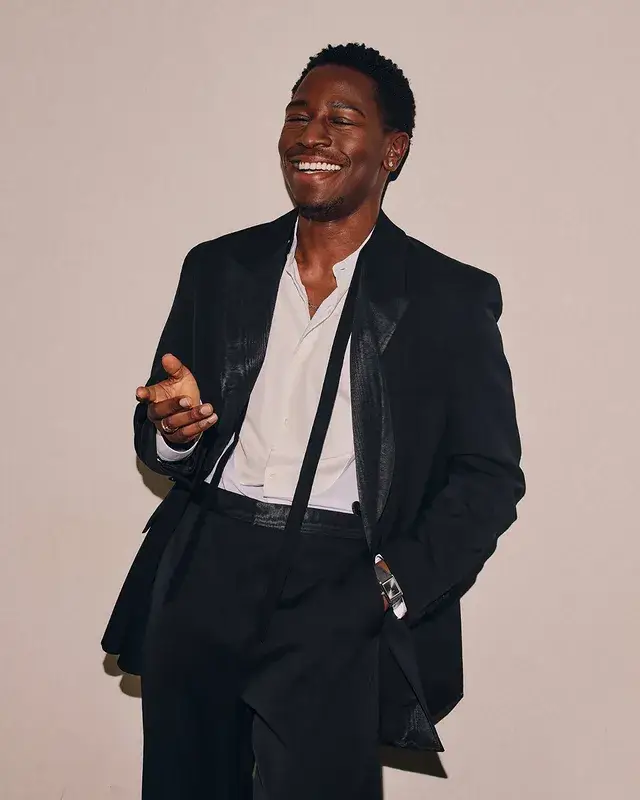 Actor David Jonsson laughs joyfully while posing in a black suit with a unbuttoned white shirt against a plain wall.