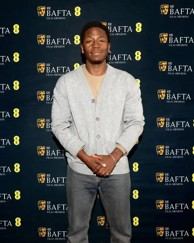 Actor David Jonsson stands with hands clasped wearing a light grey cardigan over a beige shirt at a EE BAFTA Film Awards event.