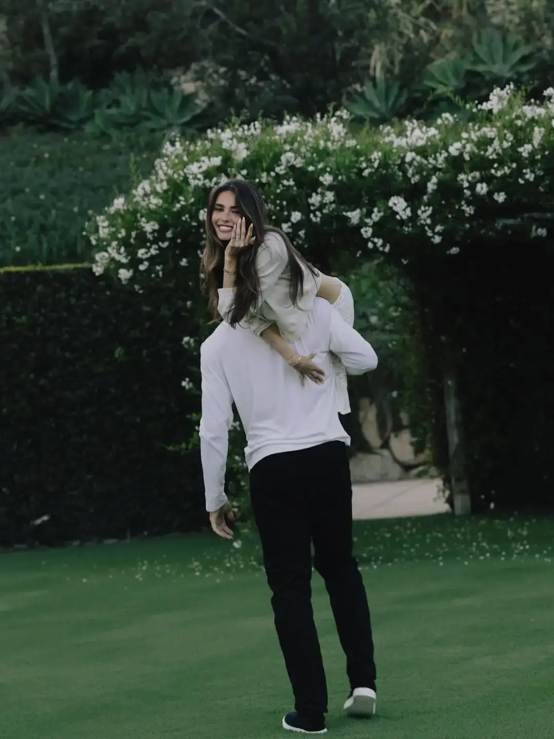 Madison Beer smiles and displays her ring while being carried over the shoulder by her fiancé Justin Herbert in a garden.
