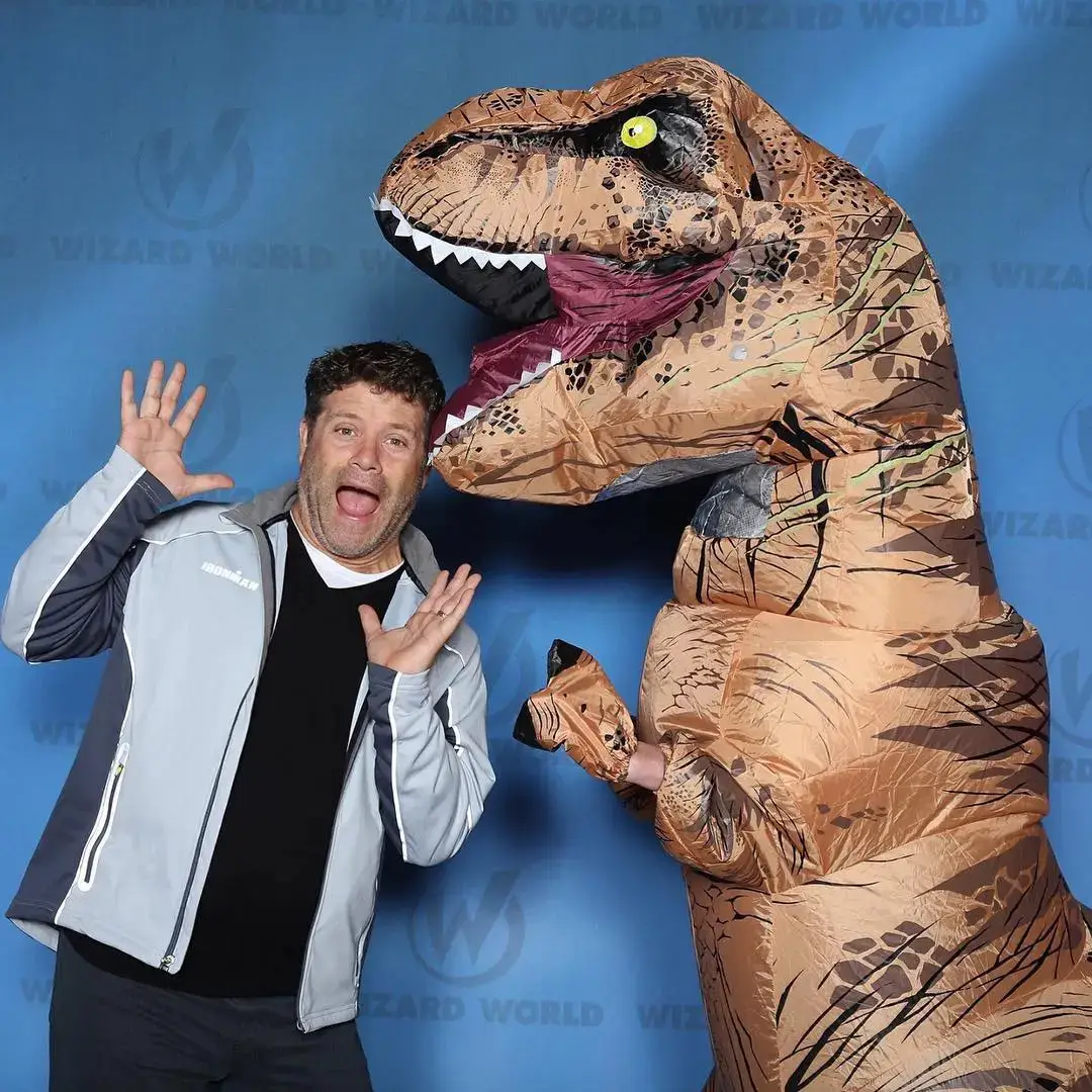 Actor Sean Astin playfully reacts with open hands alongside a person in an inflatable T-Rex suit at a Wizard World convention backdrop.