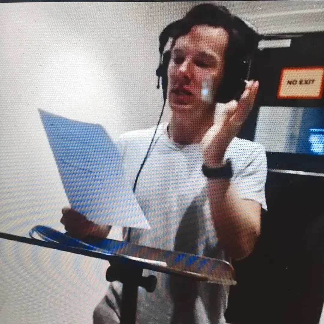 Actor Benedict Cumberbatch wearing headphones reads from a script sheet in a recording studio booth.
