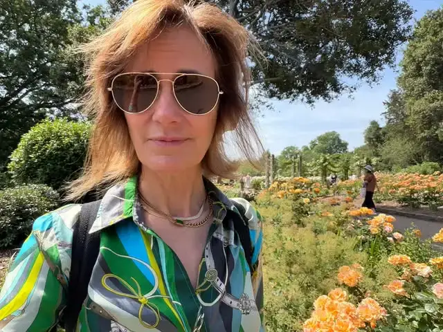 Diane Farr taking a outdoor selfie in aviator sunglasses and a colorful patterned shirt next to a blooming flower garden.