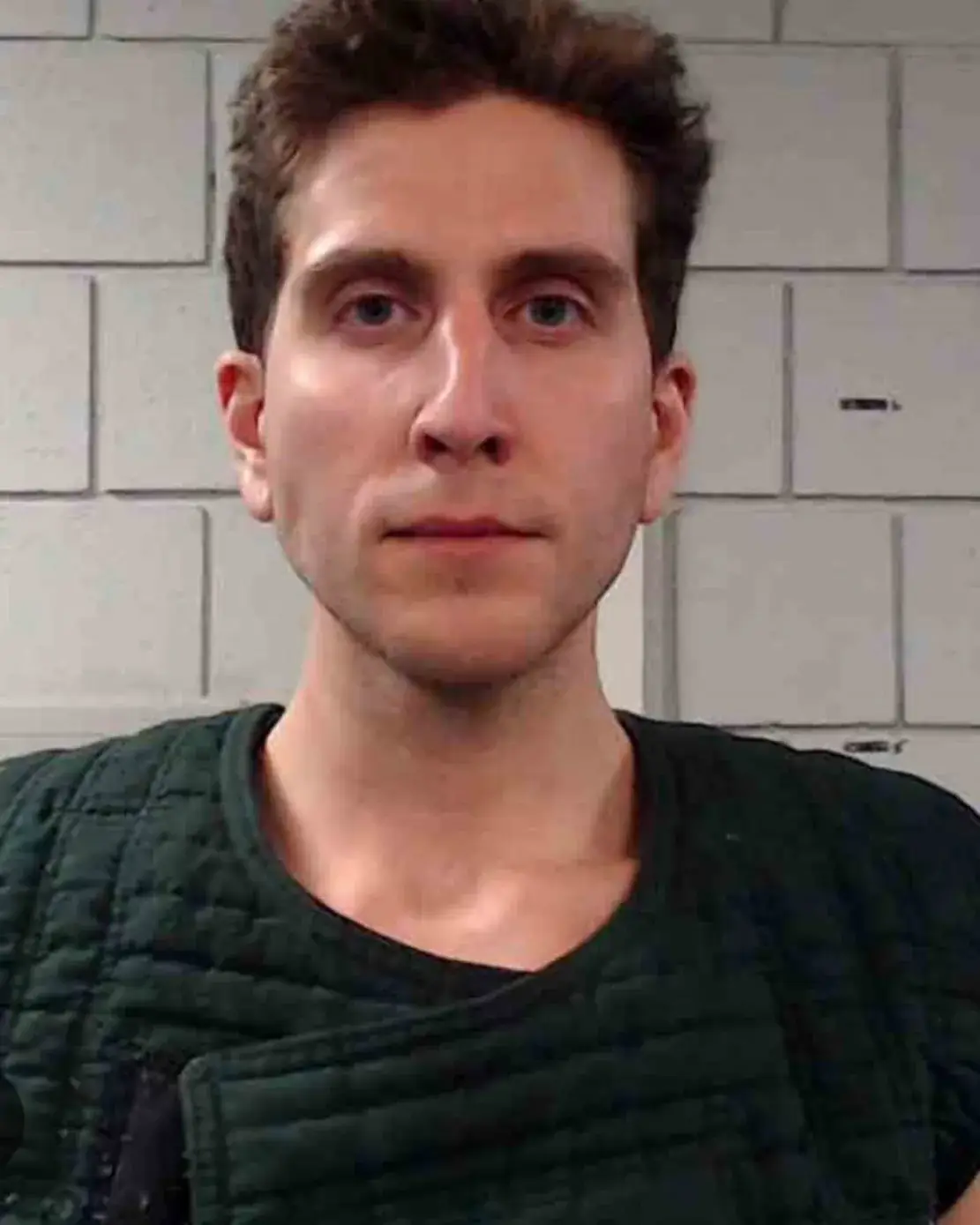 Bryan Kohberger mugshot portrait facing forward in a dark green padded vest against a cinder block wall.