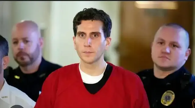 Bryan Kohberger wearing a red jail jumpsuit while flanked by law enforcement officers inside a courtroom.