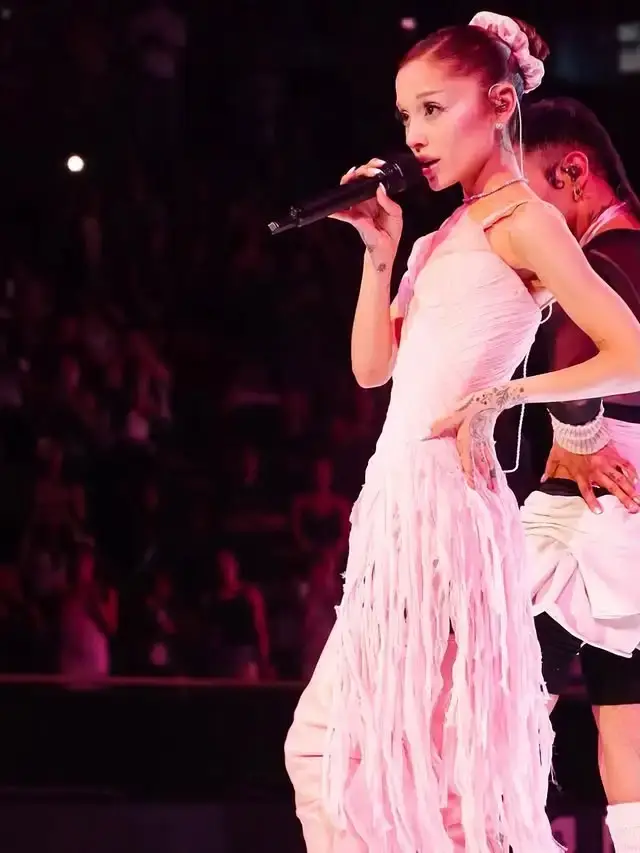 Ariana Grande singing into a microphone on stage wearing a fringed light pink dress and a high hair bun.