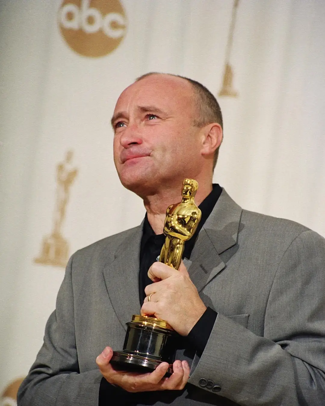 Phil Collins holding an Oscar statuette in a grey suit while looking up with an emotional smile.