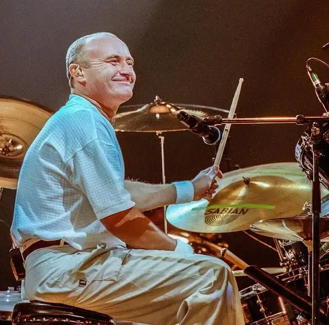 Phil Collins smiling while holding drumsticks behind a drum set during a live stage concert.
