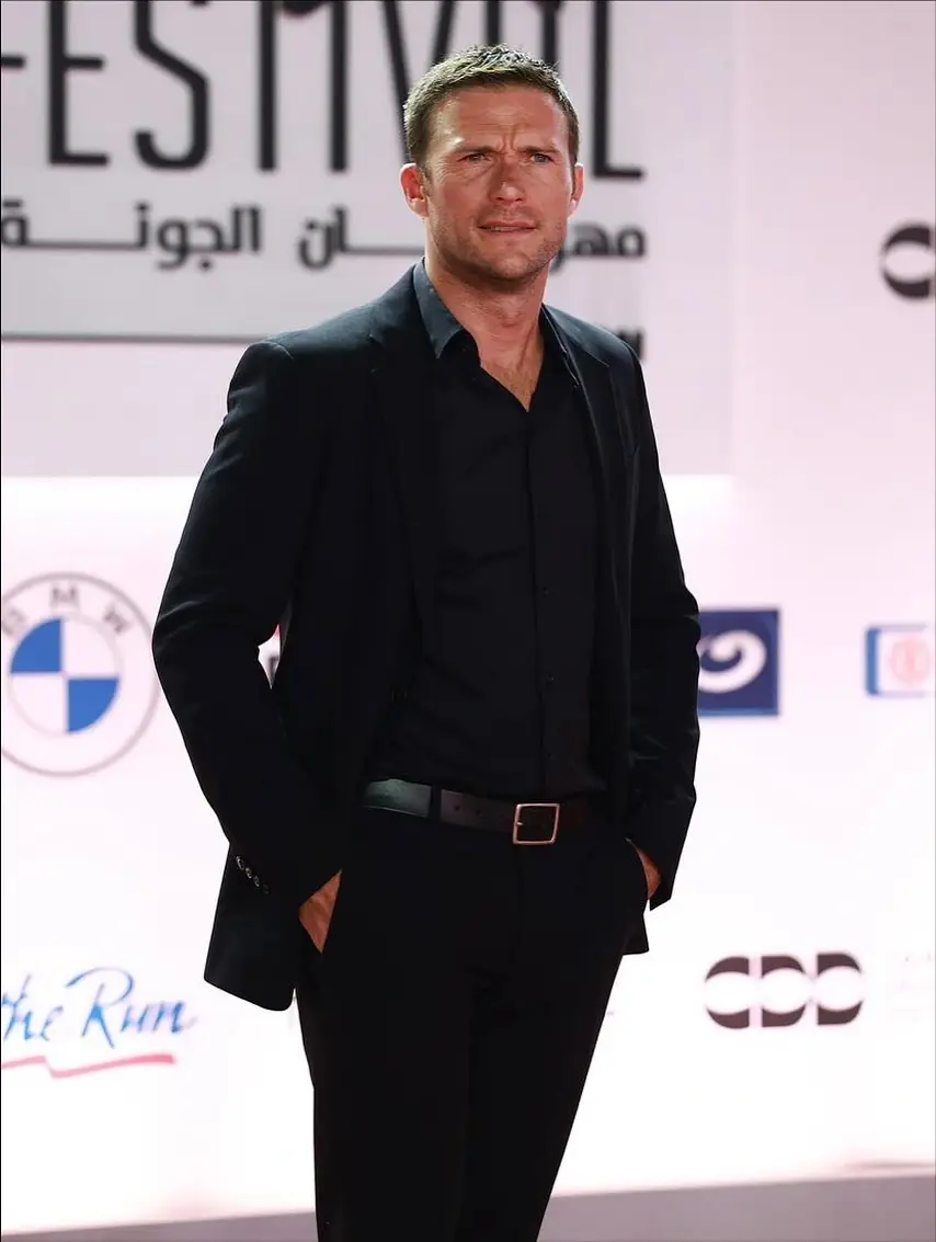 Scott Eastwood wearing an all-black suit with his hands in his pockets on a red carpet.