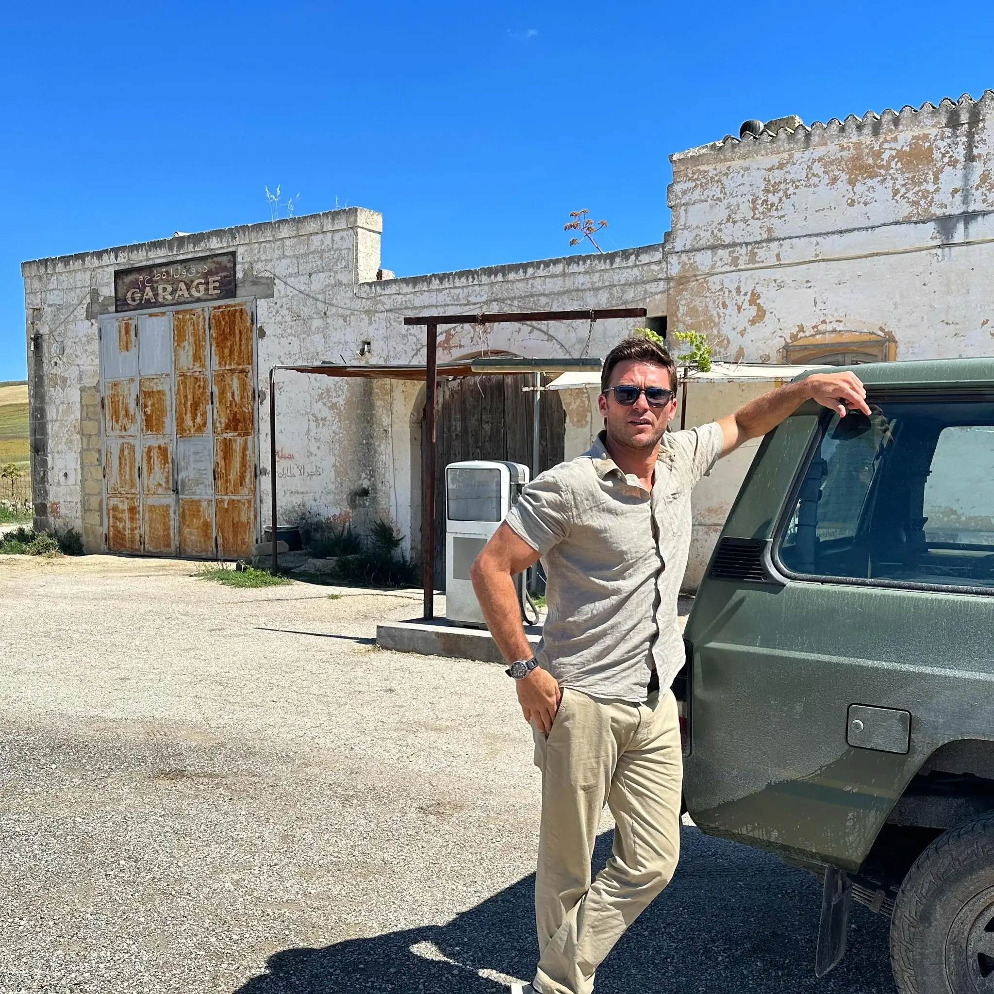 Scott Eastwood wearing sunglasses leaning against an off-road vehicle in front of an old rustic garage.