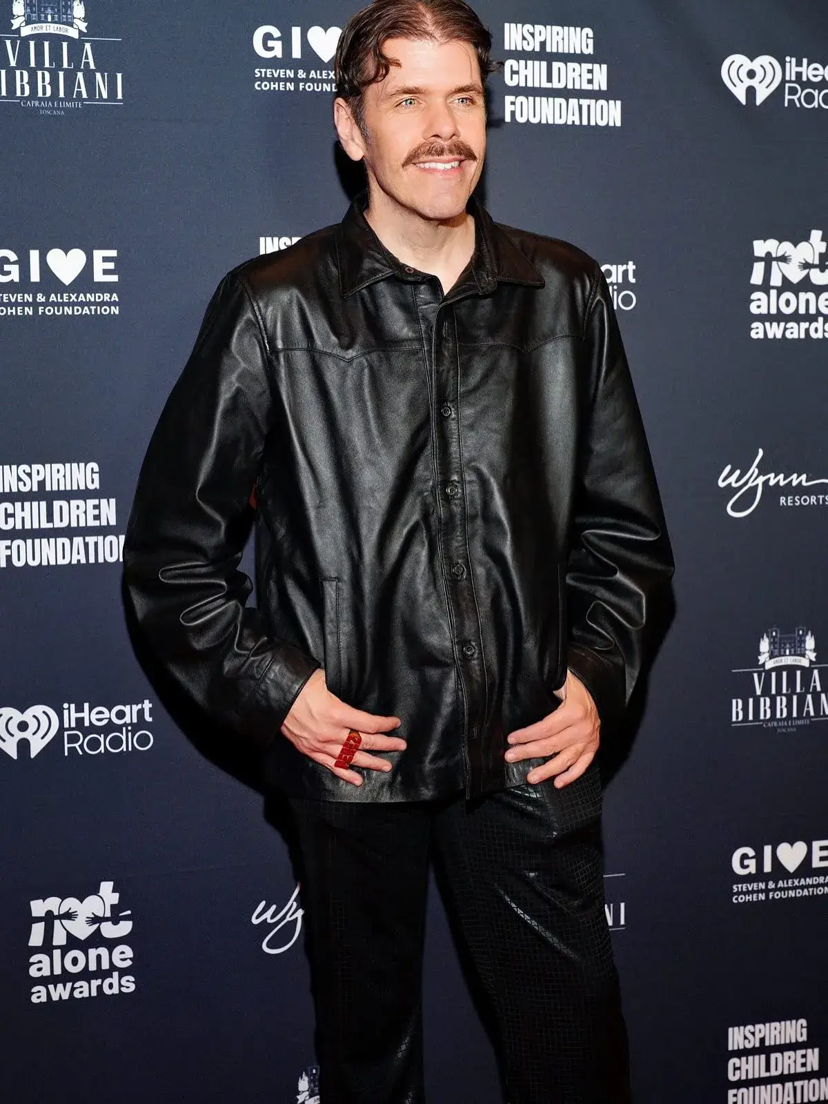 Perez Hilton sporting a mustache and a black leather jacket while posing on a red carpet backdrop.