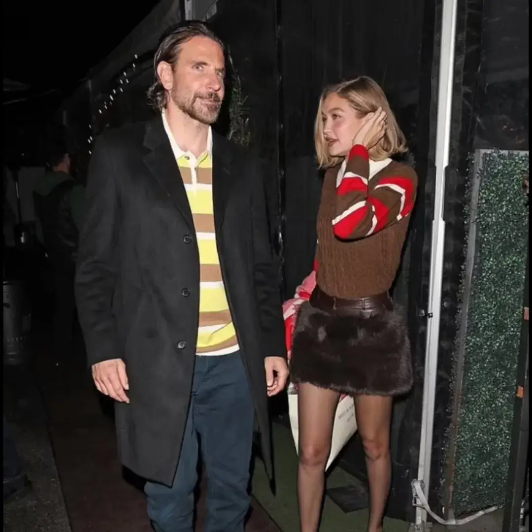 Bradley Cooper in a black coat and striped polo shirt walks alongside Gigi Hadid in a sweater and fur skirt.