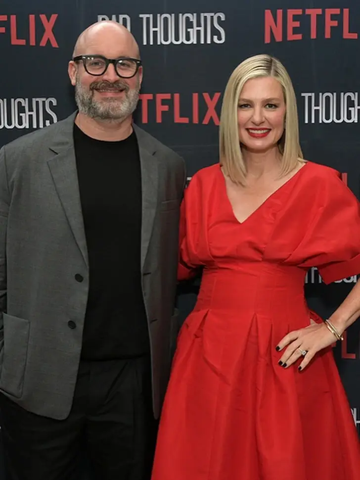 Tom Segura in a grey blazer stands alongside his ex Christina Pazsitzky in a red dress in front of a Netflix backdrop.