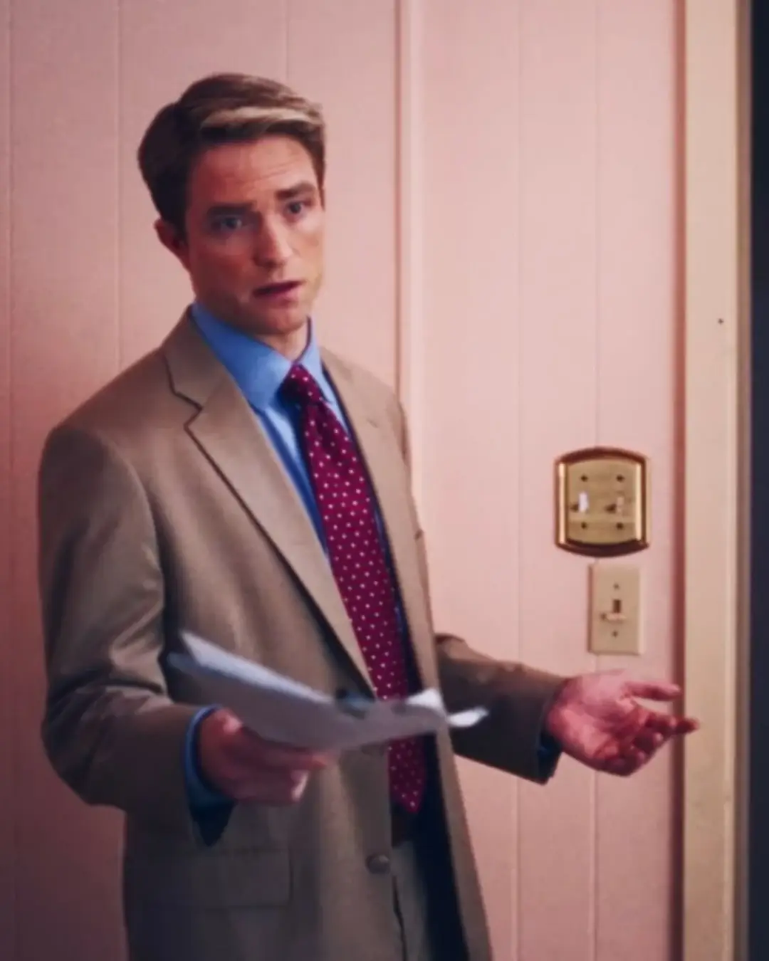 Robert Pattinson wearing a beige suit, blue shirt, and red polka-dot tie while gesturing with papers as Chris Hansen in the film Primetime.