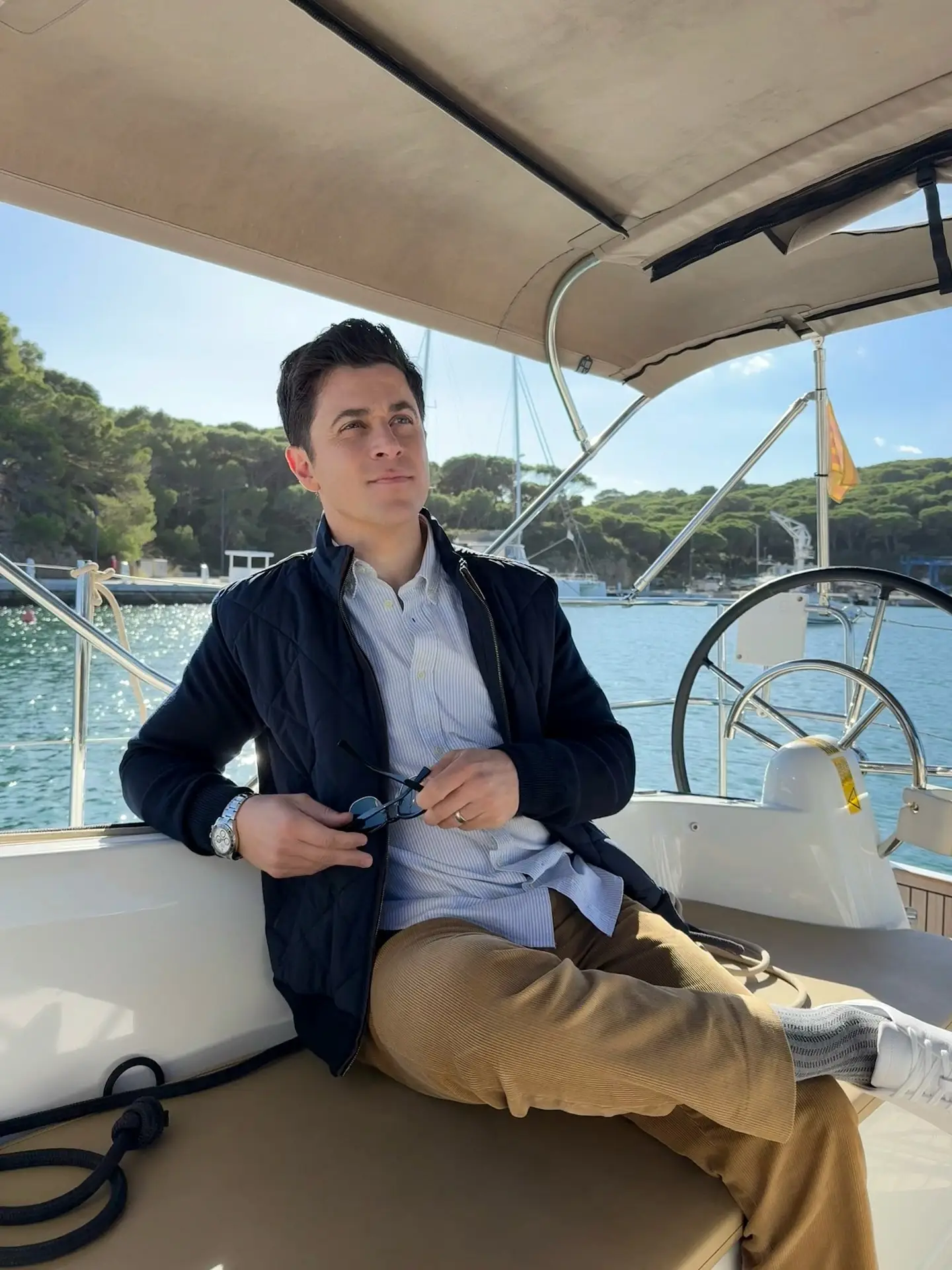 Actor David Henrie holding sunglasses while relaxing on a boat near a sunny shoreline.