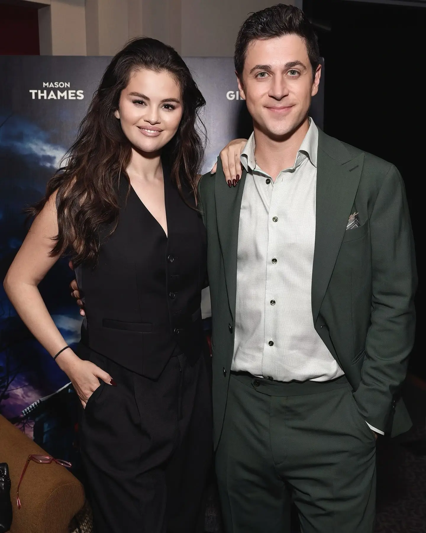 Selena Gomez in a black vest and trousers posing alongside David Henrie in a dark green suit at a red carpet premiere event.