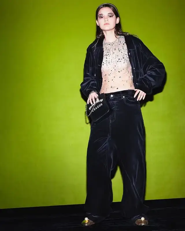 Ciara Bravo poses in a sheer embellished top, black jacket, and loose black pants against a bright green background.