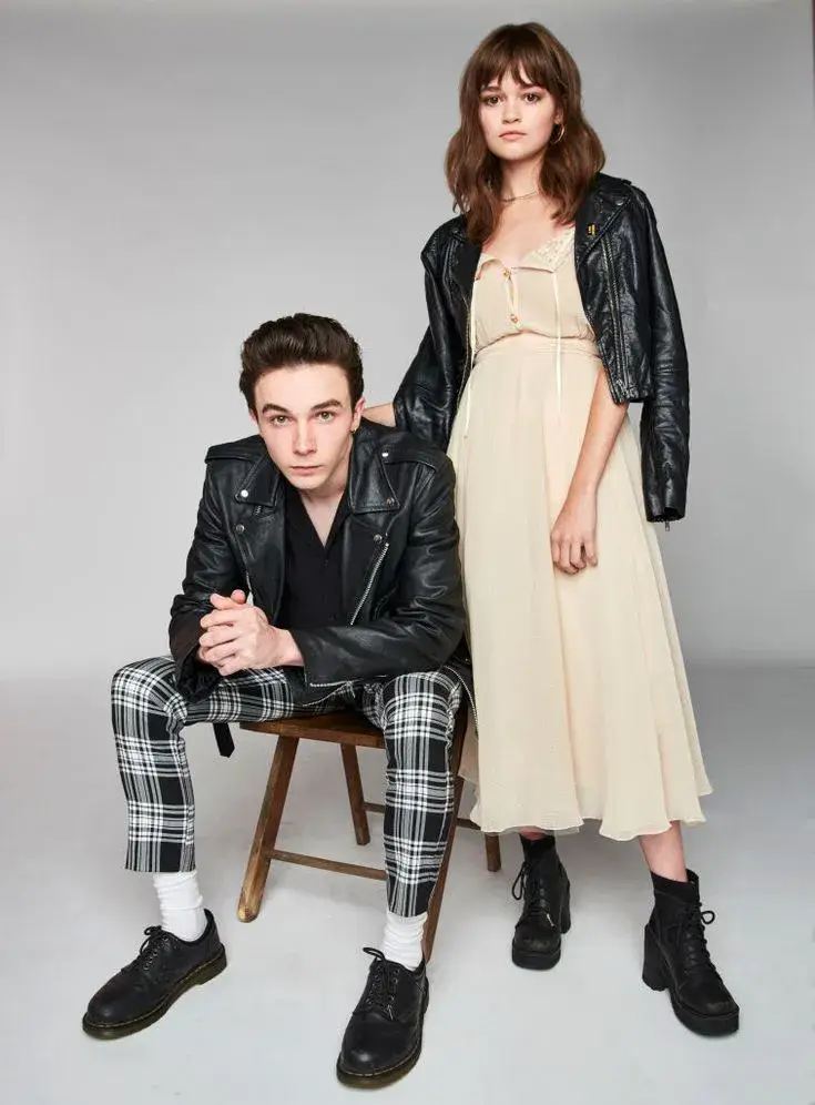 Mark McKenna seated in a leather jacket and plaid pants beside Ciara Bravo in a cream dress and black jacket.