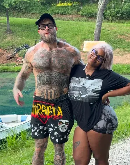 Shirtless tattooed man Dalton posing outdoors near water with Sako wearing a black graphic t-shirt and glasses.