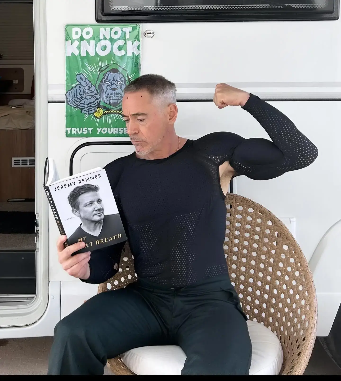 Robert Downey Jr. flexing his bicep while reading Jeremy Renner’s memoir in a tight black top outside a trailer, putting on a humorous body show.