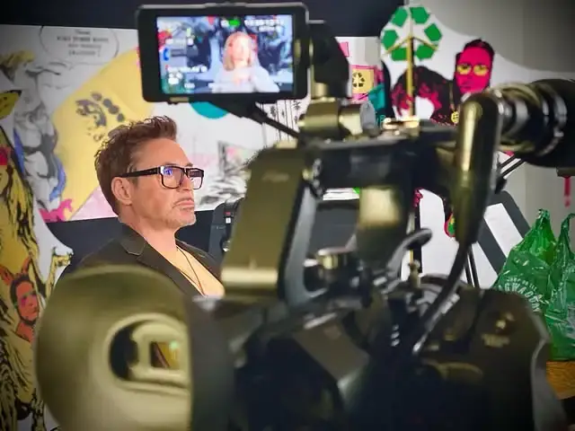 Robert Downey Jr. wearing glasses looks thoughtfully off-camera on a production set behind a large video camera.