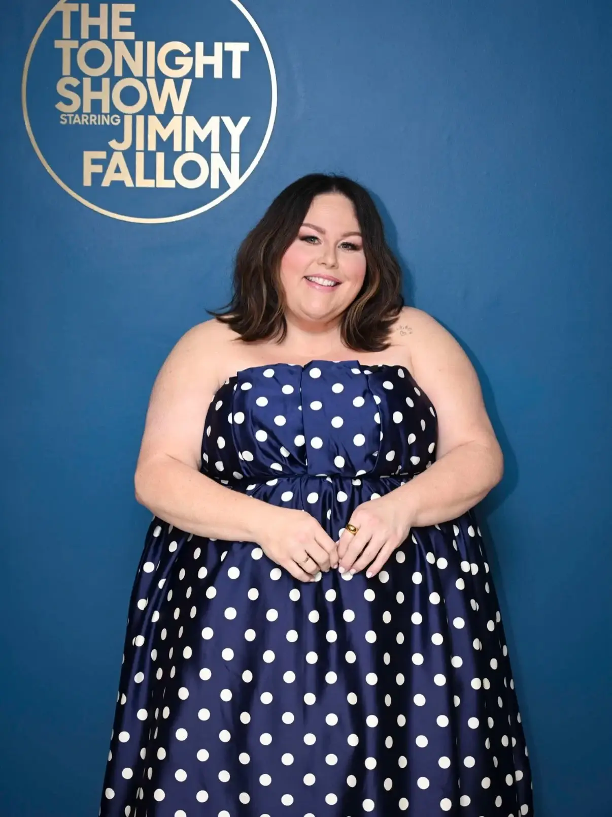 Chrissy Metz smiling in a strapless blue and white polka-dot dress in front of The Tonight Show Starring Jimmy Fallon backdrop.