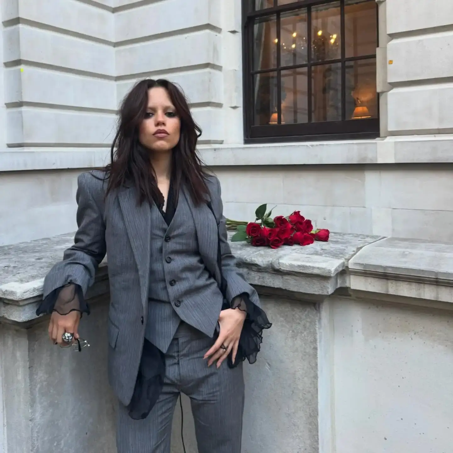 Jenna Ortega poses in a tailored grey pinstripe suit with ruffled cuffs near a bouquet of red roses.
