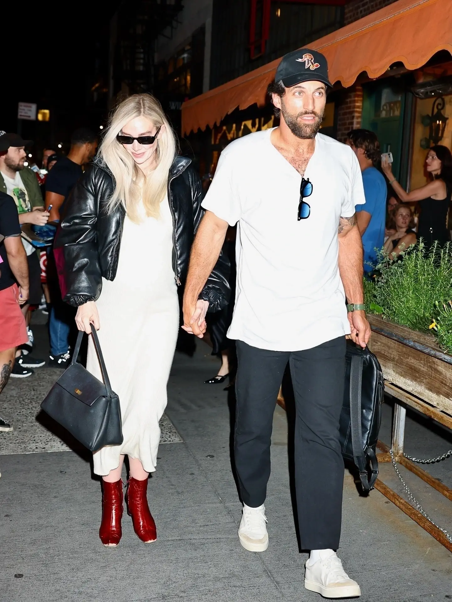 Vanessa Kirby wearing a black leather jacket and white dress holding hands with her fiancé Paul Rabil on a nighttime street walk.