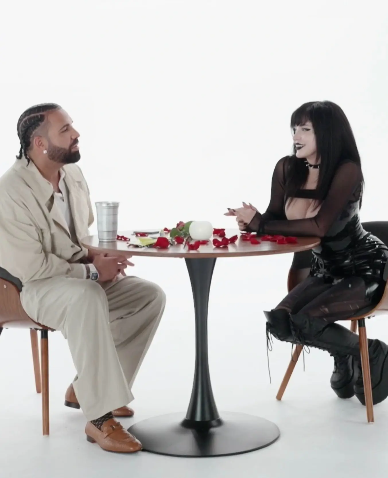 Drake in a cream suit sitting across a small round table with rose petals from Pinkchyu in a black gothic outfit during a date.