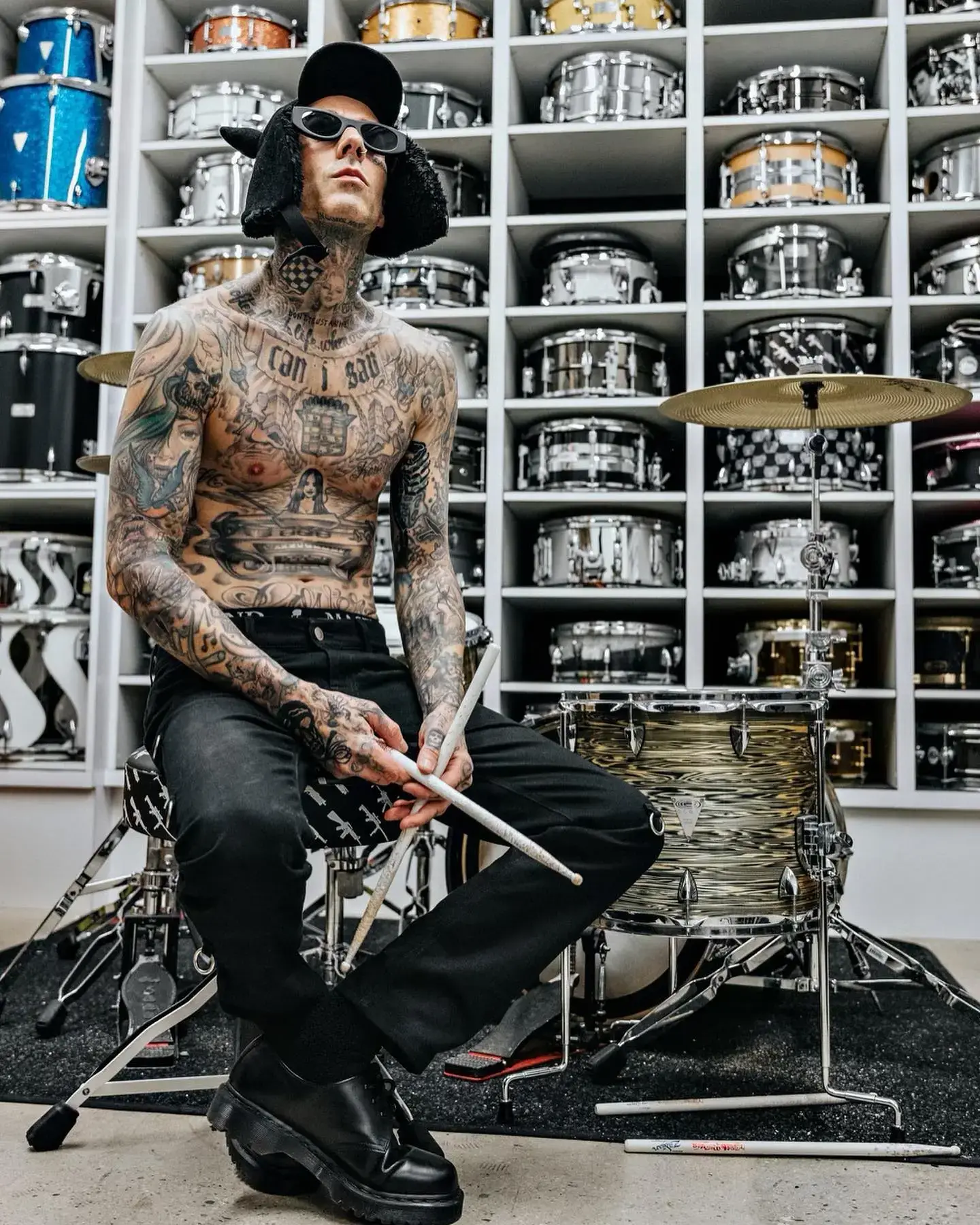 A shirtless, heavily tattooed Travis Barker sits holding drumsticks behind a drum kit in front of shelves stocked with snare drums.