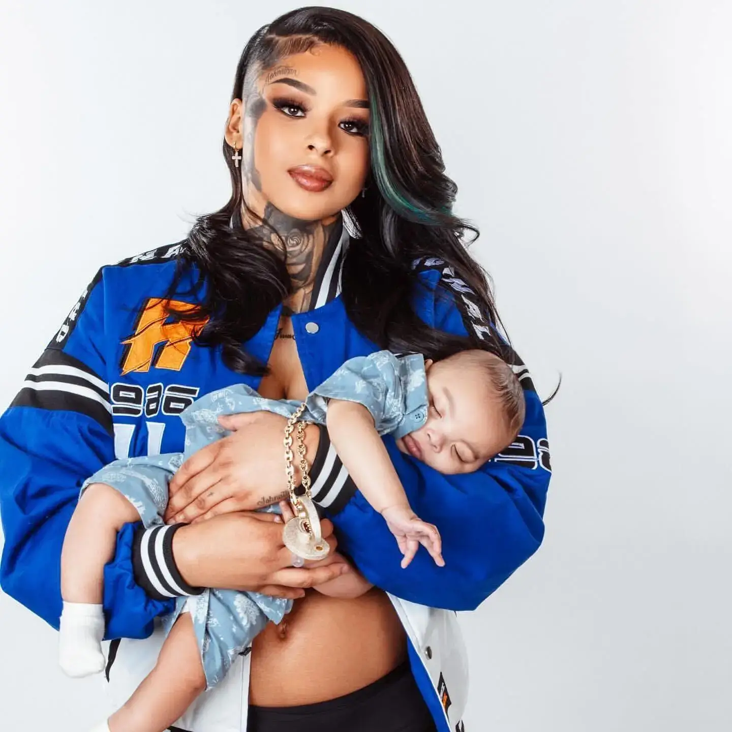 ChriseanRock in a blue racing jacket cradles her sleeping baby boy, Chrisean Malone Jr., in her arms against a white background.