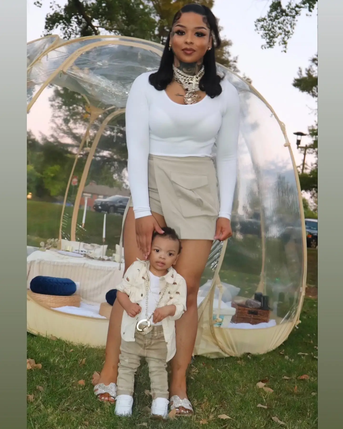 Chrisean Rock stands outdoors in front of a clear bubble tent with her son Chrisean Jesus posing in front of her.