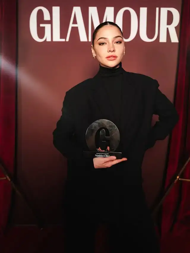 Madelyn Cline dressed in a chic all-black turtleneck and blazer holding a Glamour award in front of a red backdrop.