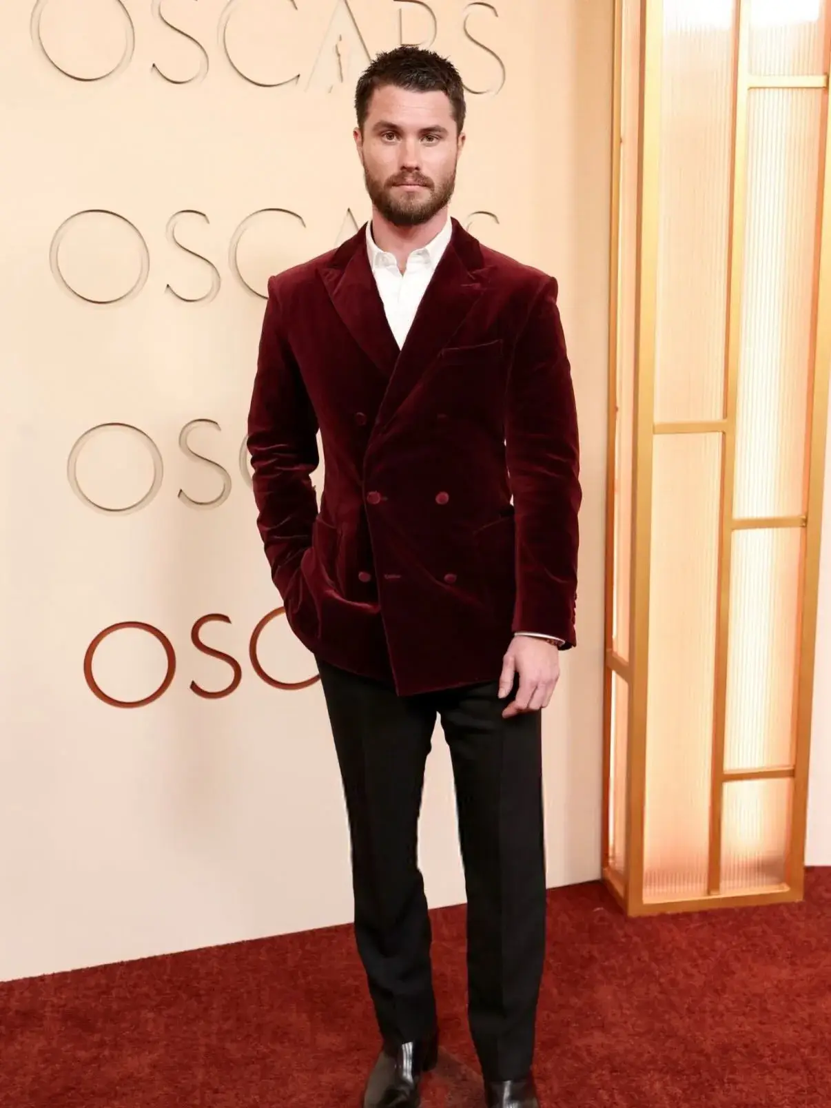 Chase Stokes posing full-length in a maroon velvet double-breasted blazer and black trousers on the Oscars red carpet`.